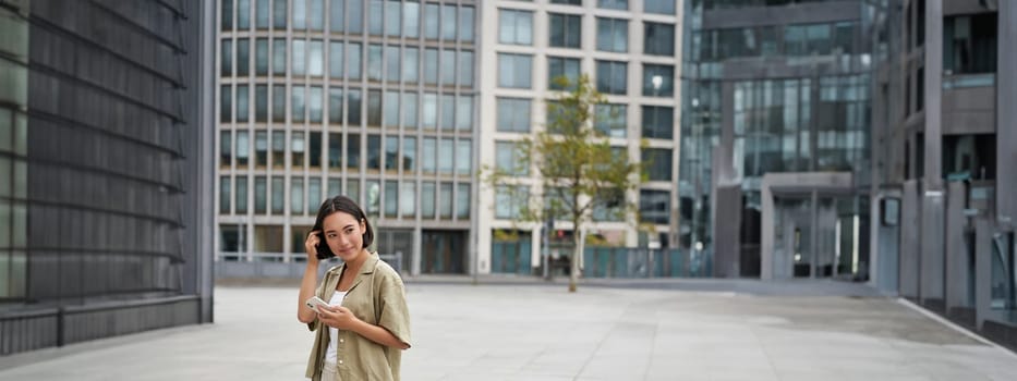 Portrait of asian woman walking in city. Street style shot of girl with smartphone, posing outdoors on street.