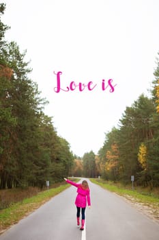 a girl in a pink raincoat and in rubber boots walks on the road in the middle of the forest-inscription love is.