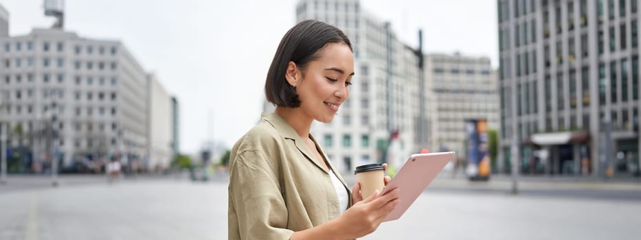 Asian girl walking on street, reading on tablet and drinking coffee outdoors.