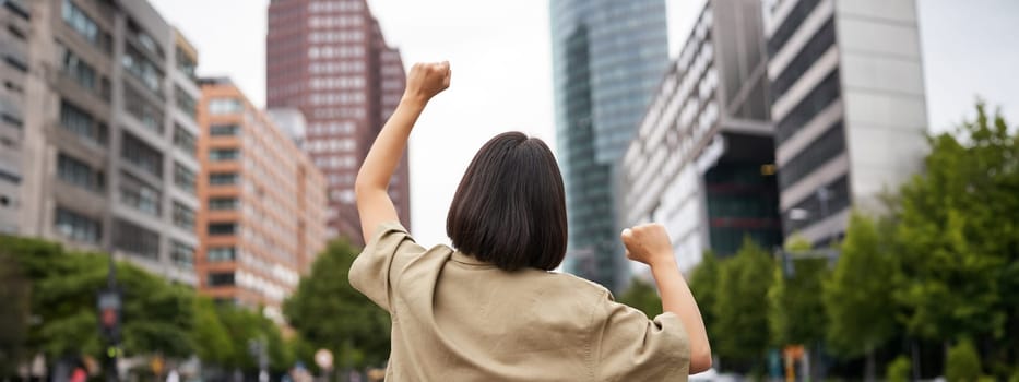 Excitement. Happy girl rear view, raising her hands up and looking at big city skyscrappers, feeling confident, cheering and triumphing.