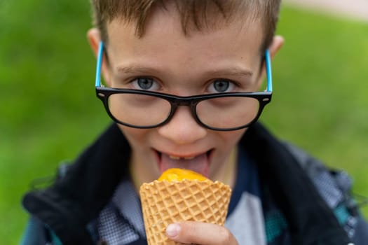 Funny Caucasian boy eats ice cream in a waffle cone on the street in the park, smiles and looks at the camera. A happy Caucasian boy with glasses walks in the park.