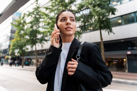 Wide portrait of siren businesswoman in black suit holding a phone to ear looking away in modern city street.