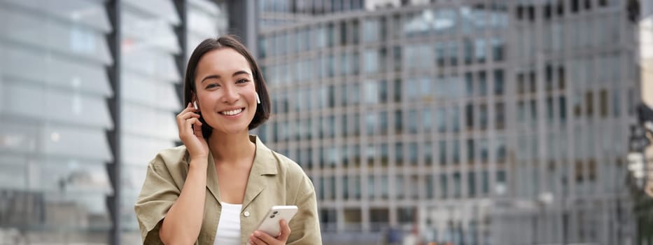 Young smiling asian woman in wireless earphones, walks on street with smartphone and headphones.