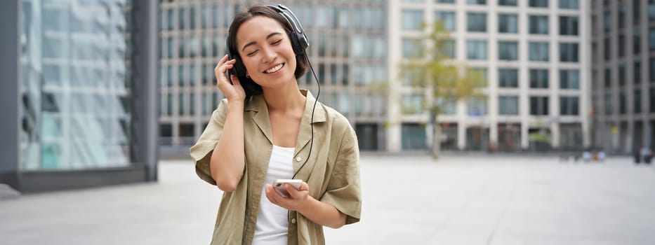 Streaming services concept. Happy asian girl listens music in headphones, holds mobile phone, choosing track or podcast, walking on street.