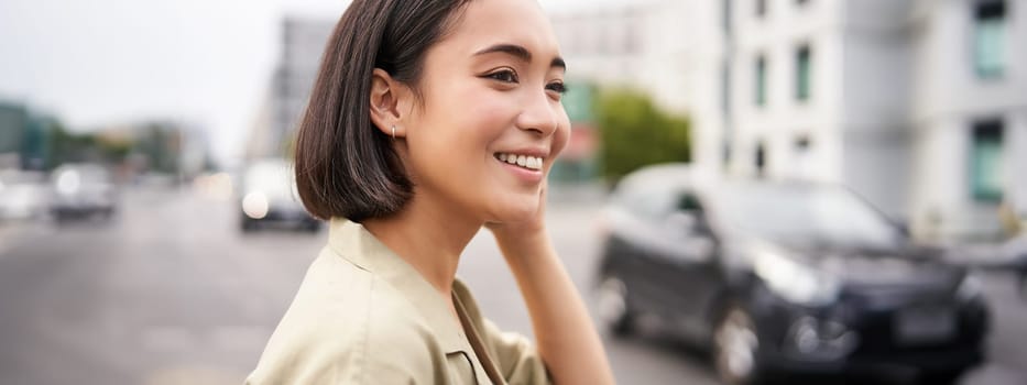 Close up portrait of beautiful young woman, asian girl stands outdoors on street, smiling happy.