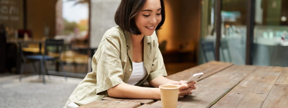 Portrait of young woman enjoying her coffee, drinking takeaway on bench in city, using smartphone. Copy space