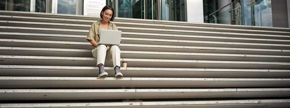 Asian girl student sits on stairs near campus, types on laptop, does her homework outdoors.