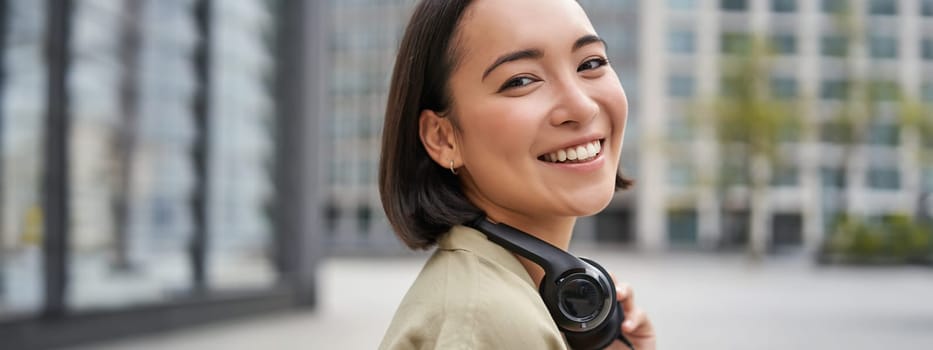 Portrait of happy asian woman in headphones, enjoys music while walking in city, smiling and laughing.