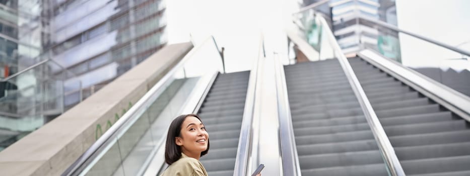 Young happy woman standing on escalator with smartphone, going up, walking in city.