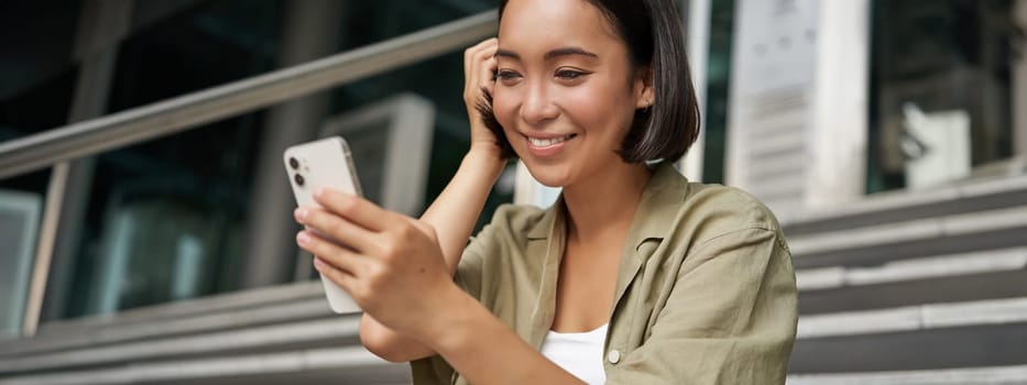 Portrait of asian girl takes selfie on mobile phone. Korean woman smiling, video chat on smartphone app while sits outside on stairs.