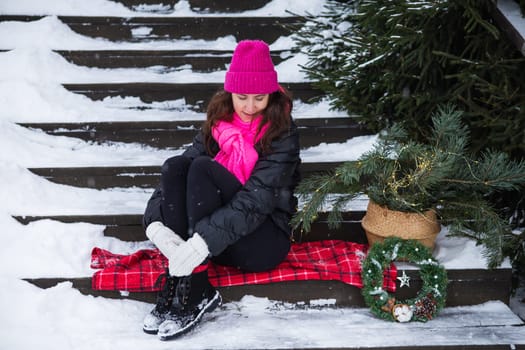 A woman sits on wooden stairs covered with snow in a pink hat near a Christmas tree in a basket, lights, gifts. New Year's magical atmosphere of coziness, love and warmth