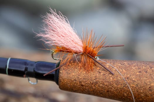 Dry fly pattern in orange with rubber legs on the cork grip of a fly fishing rod at the river.