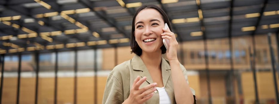 Young asian woman smiles, talks on mobile phone and walks in city centre, poses outdoors.