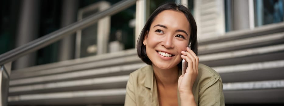 Portrait of asian girl smiles while talks on mobile phone. Young woman calling a friend, sitting on stairs. Technology concept