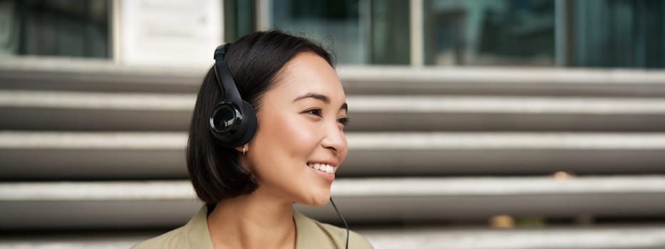 Smiling asian girl, laughing, listening music in headphones, sitting outdoors. Uni student enjoying free time.