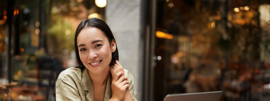 Vertical shot of young asian woman working on remote from outdoor cafe, sitting with laptop and smiling, studying.