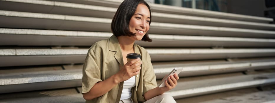 Portrait of asian woman with smartphone, drinks coffee and watches videos on mobile phone. Girl with telephone sits on stairs outdoors.