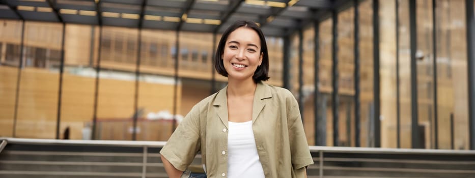 Portrait of young confident female model, girl in casual clothes, posing outside near glass building, smiling at camera.