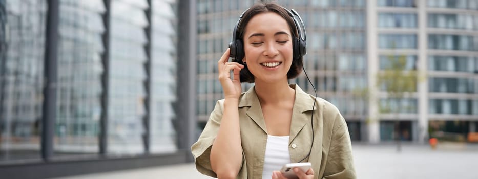 Streaming services concept. Happy asian girl listens music in headphones, holds mobile phone, choosing track or podcast, walking on street.