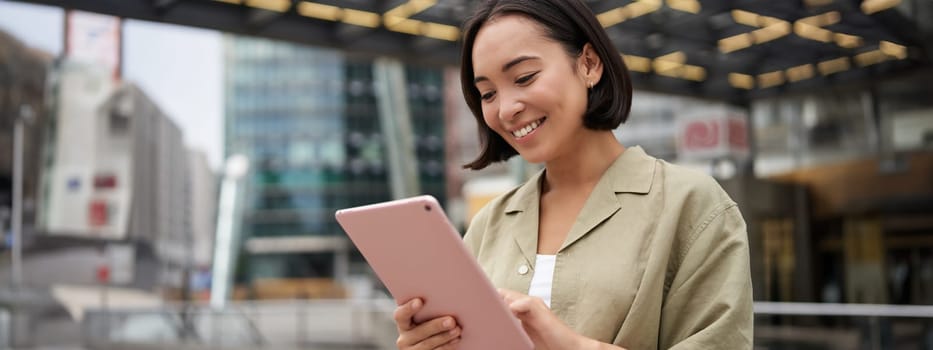 Portrait of asian woman standing on street, using tablet, smiling with carefree face expression, outdoor shot.