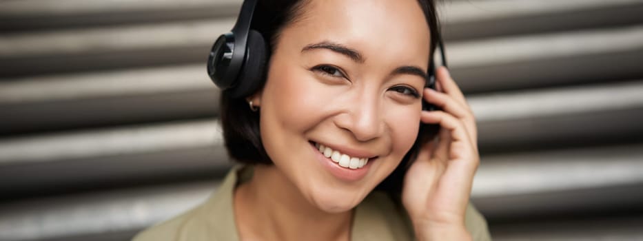 Close up portrait of smiling asian girl in headphones, listens to music outdoors, looking happy. People concept