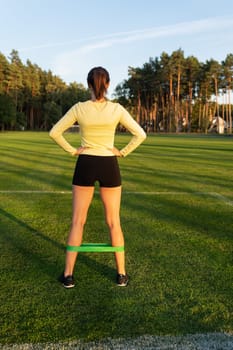 A girl stands in shorts at the stadium and does exercises with a sports elastic band. Sports and health concept