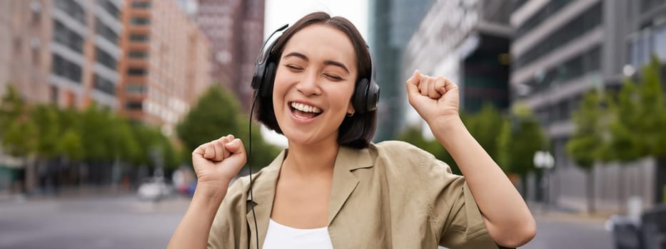 Dancing girl feeling happy in city. Asian woman dancing and listening music in headphones, posing on street.