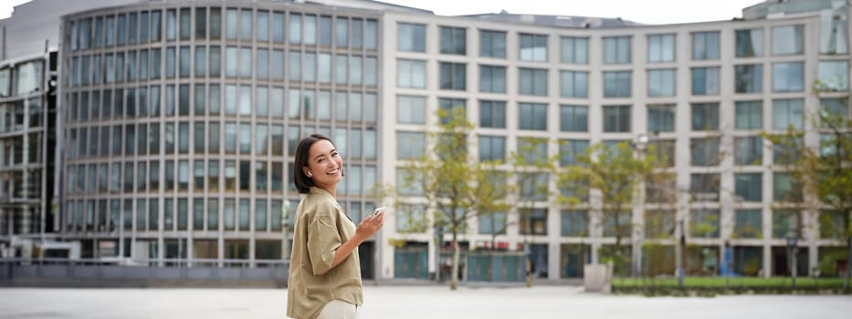 Modern people. Young woman walks on street of city and listens music in wireless headphones, holds smartphone.