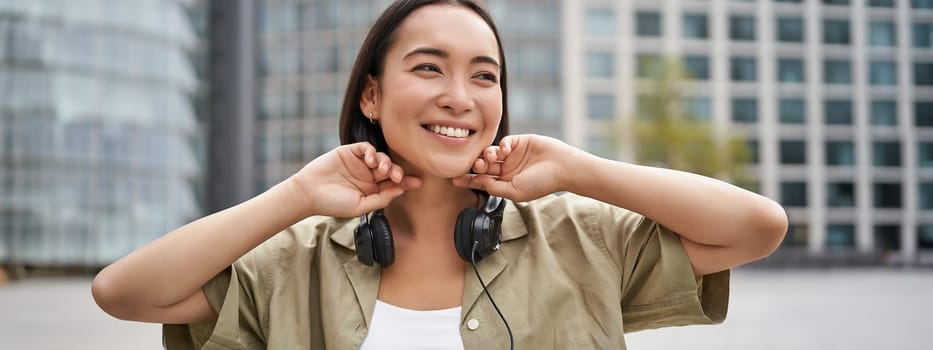 Portrait of smiling asian girl with headphones, posing in city centre, listening music.