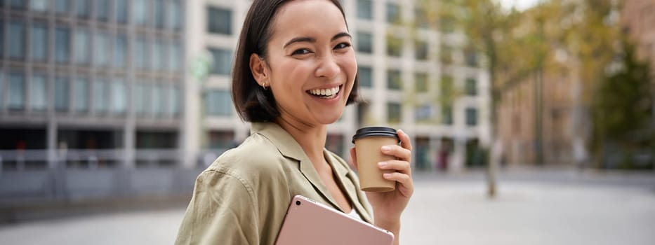 Portrait of asian girl with tablet, drinks coffee on street, walking in city centre.