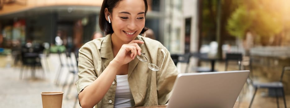 Smiling young korean woman looks at her laptop screen with pleased face, works remotely from outdoors, drinks coffee.