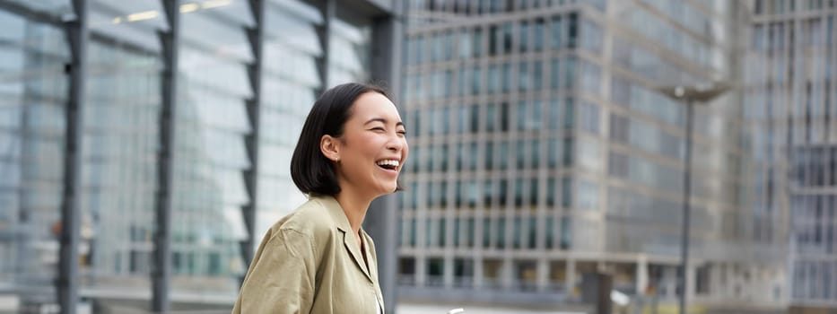 Vertical shot of beautiful asian woman walking on street, laughing and looking happy, enjoying the day.