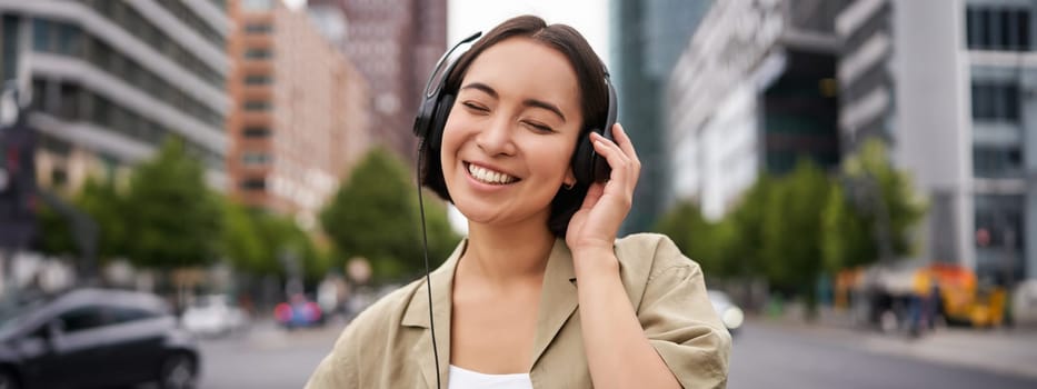 Portrait of smiling asian woman in headphones, standing in city centre on street, looking happy, listening to music.