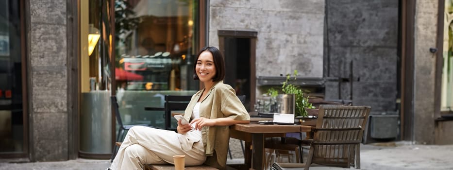 Portrait of stylish young korean woman sits in cafe, holds smartphone, smiles, enjoys coffee outdoors.