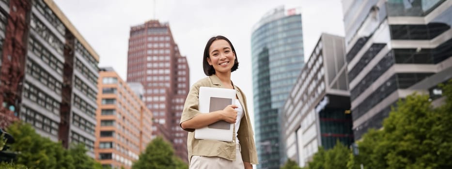 Portrait of young asian woman, looking happy and confident, going to work or university, city skyscrappers behind her, holding laptop and notebook.