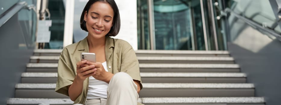 Smiling asian girl sits on stairs near building entrance, using mobile phone app. Happy young woman rests with smartphone in her hands outdoors.