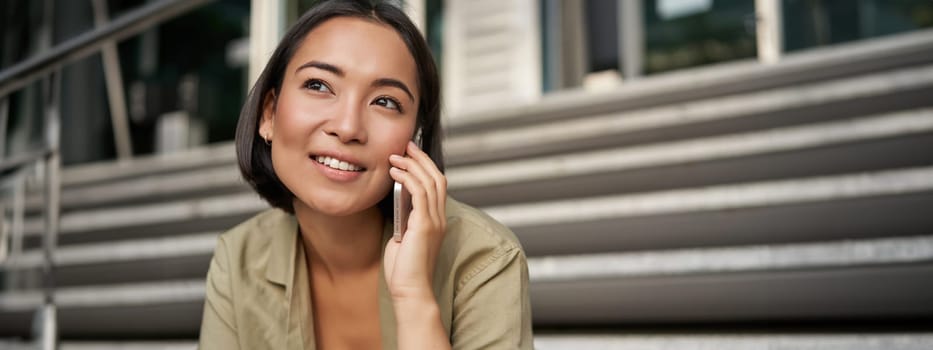 Cellular technology. Smiling asian girl talks on mobile phone, makes a call. Young woman with telephone sits on stairs.