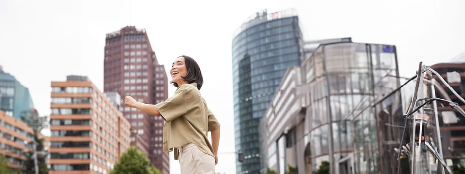 Vertical shot of young asian woman posing happy, raising hands up and dancing, triumphing, celebrating victory, enjoying day out in city.