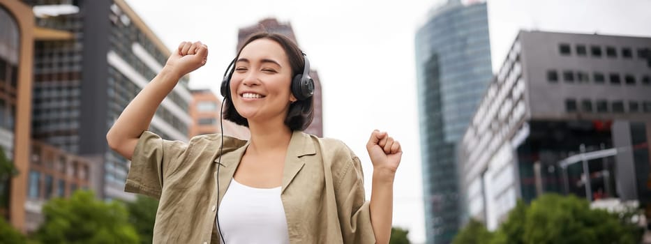 Happy asian woman in headphones, listening music and dancing on street of city centre, smiling with hands up.