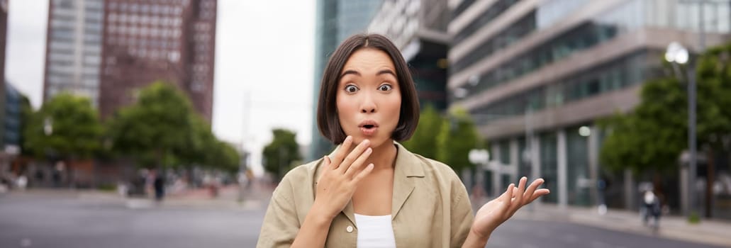 Modern asian girl standing on street with surprised face, looking impressed and amazed, hear awesome news.