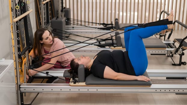 Overweight caucasian woman doing pilates exercises on reformer with personal trainer