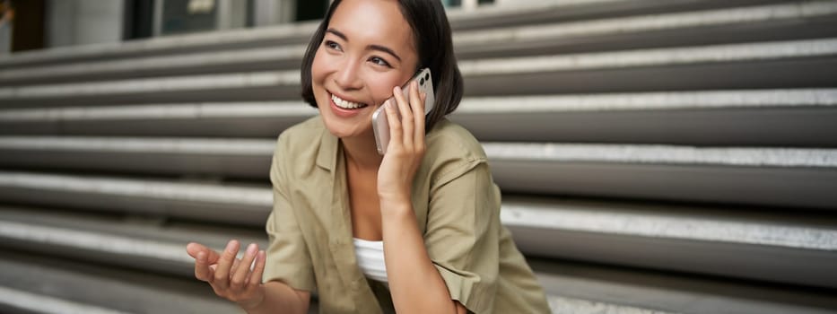 Close up portrait of smiling asian girl talks on mobile phone, sits outside on street stairs. Young woman calling friend on smartphone.