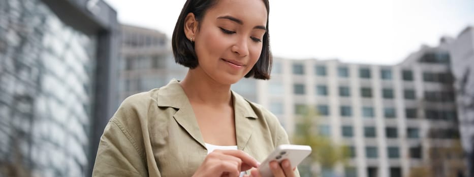 Mobile technology. Smiling asian woman using smartphone app, looking at her telephone on street, checking map, calling or texting someone.