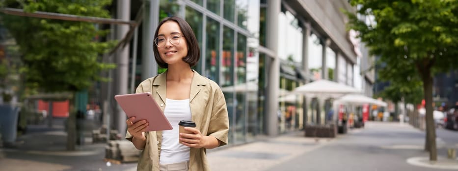 Portrait of smiling young woman walking in city with tablet, drinking takeaway coffee, going down the street with happy expression.