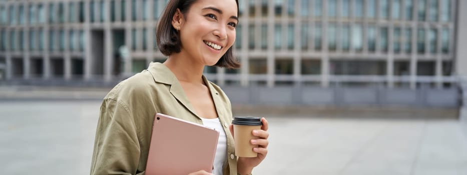 Portrait of happy asian girl with tablet and coffee, walking on street with pleased smile.