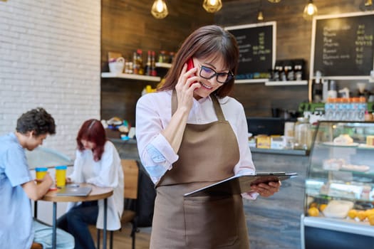Portrait of confident middle aged woman coffee shop worker. Female in apron holding clipboard with orders talking on cell phone with cafeteria customers. Small business work people concept