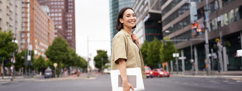 Outdoor shot of asian girl with laptop, going somewhere in city centre, walking on street, going to work.