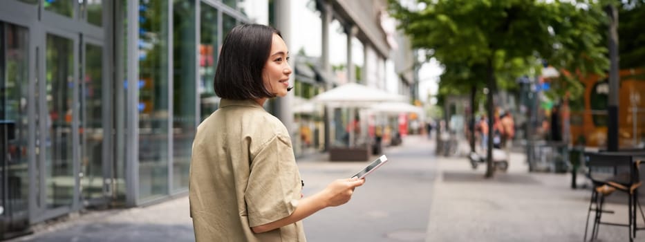 Rear shot of young woman walking in city, going down the street and smiling, holding smartphone. View from behind.