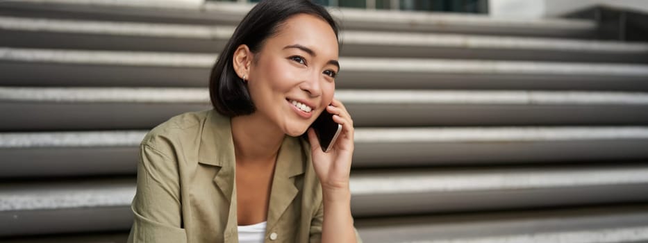 Portrait of beautiful asian girl talks on mobile phone, sits on street stairs. Woman with smartphone smiling, making a call.