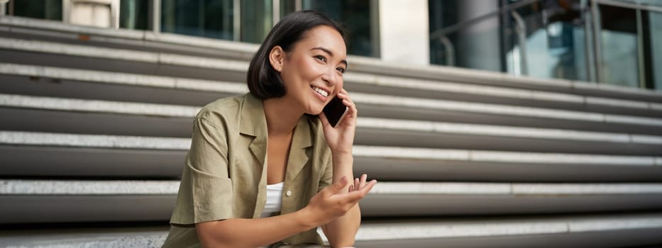 Portrait of beautiful asian girl talks on mobile phone, sits on street stairs. Woman with smartphone smiling, making a call.